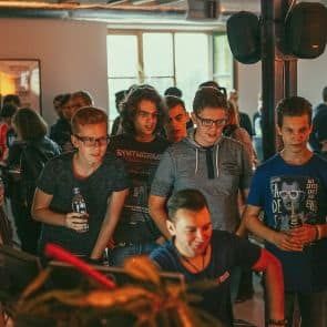 Jaunu vīriešu grupa, daži turot rokās dzērienus, vērīgi skatās uz datora ekrānu vai spēļu konsoli telpā, kas piepildīta ar citiem cilvēkiem. Atmosfēra šķiet dzīva un sabiedriska.